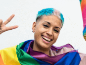 A smiling racialized queer person holding a rianbow coloured megaphone in one hand, and making a peace sign in the other hand, wearing the PRide glad around her shoulder.