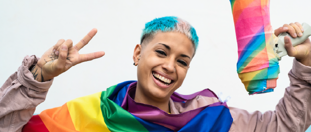 A smiling racialized queer person holding a rianbow coloured megaphone in one hand, and making a peace sign in the other hand, wearing the PRide glad around her shoulder.